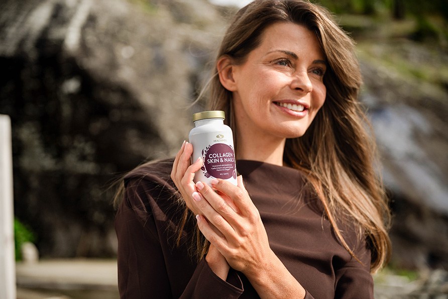 En kvinde med langt brunt hår og brun trøje står foran en klippe og holder en bøtte Healthwell Collagen Skin & Nails.