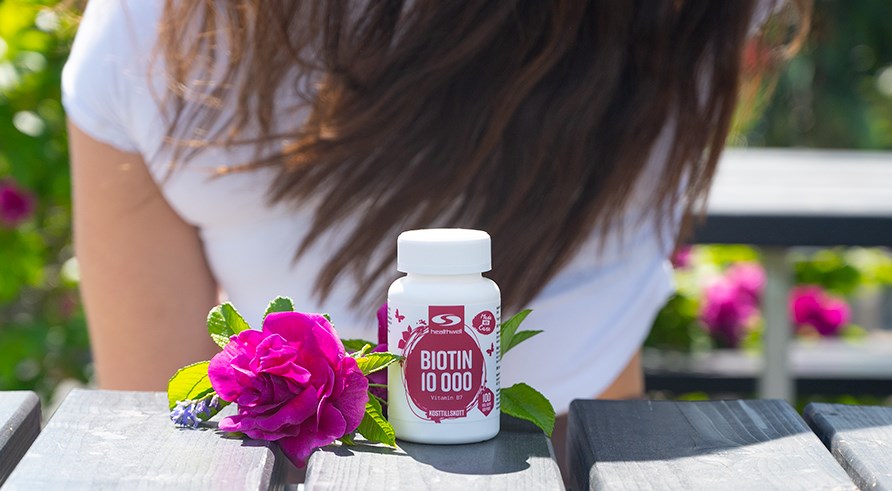 En btte Healthwell Biotin 10 000 str p en bnk ved siden af en lyserd blomst.