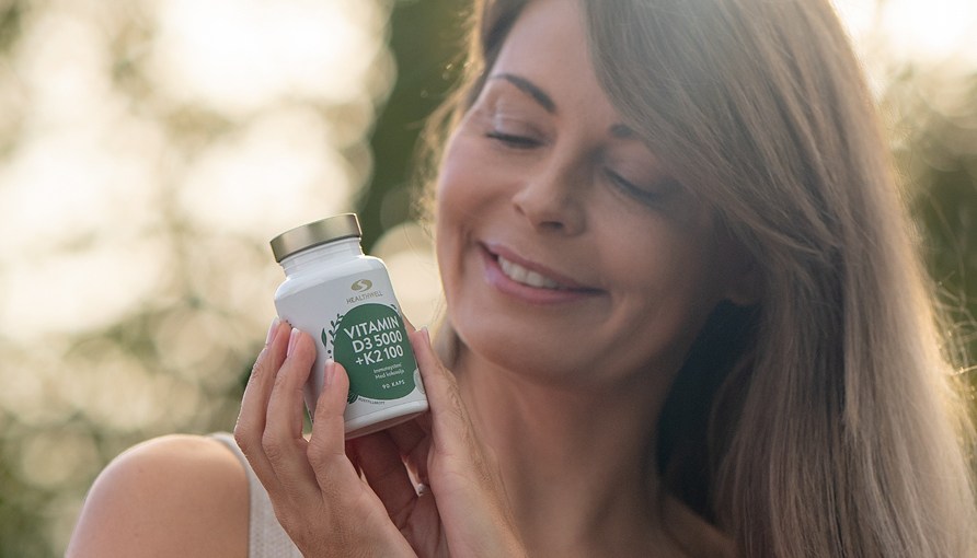 Glad kvinde holder Healthwell Vitamin D3 5000 + K2 100 ude i naturen.