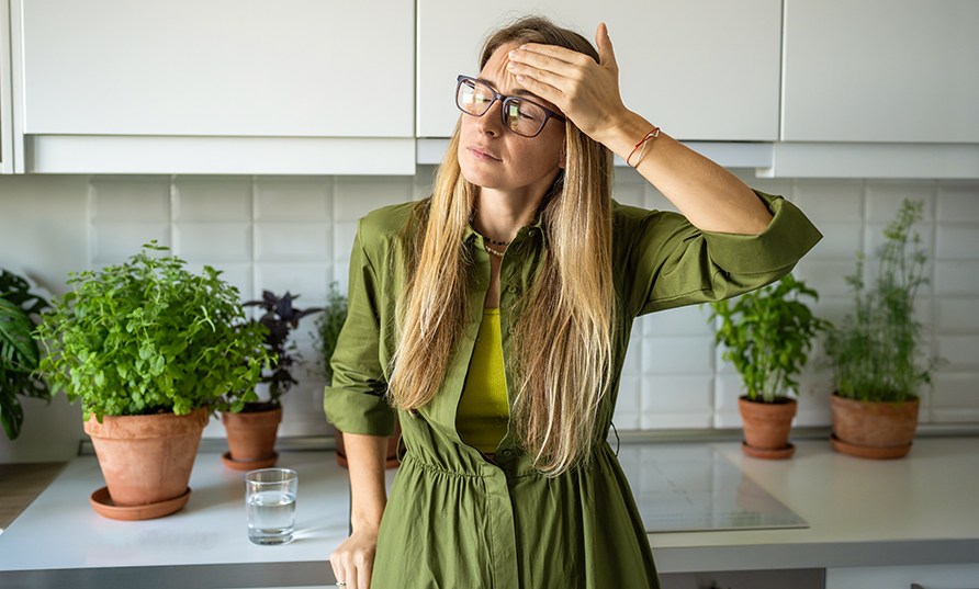 En kvinde str i kkkenet foran grnne bladplanter med hnden for panden.