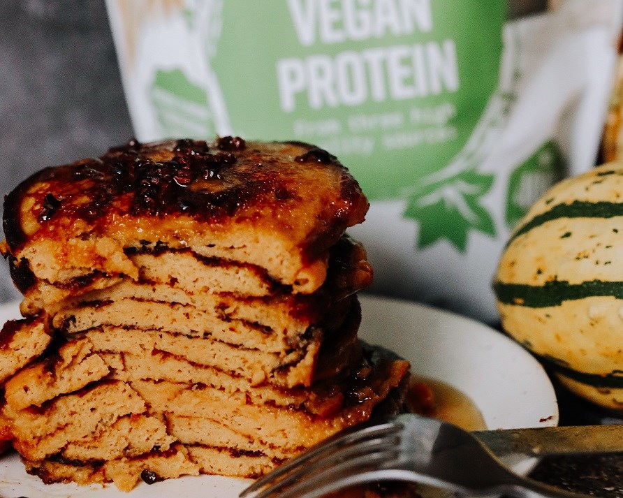 En stak proteinpandekager med grskar str foran en pose Healthwell Vegan Protein.