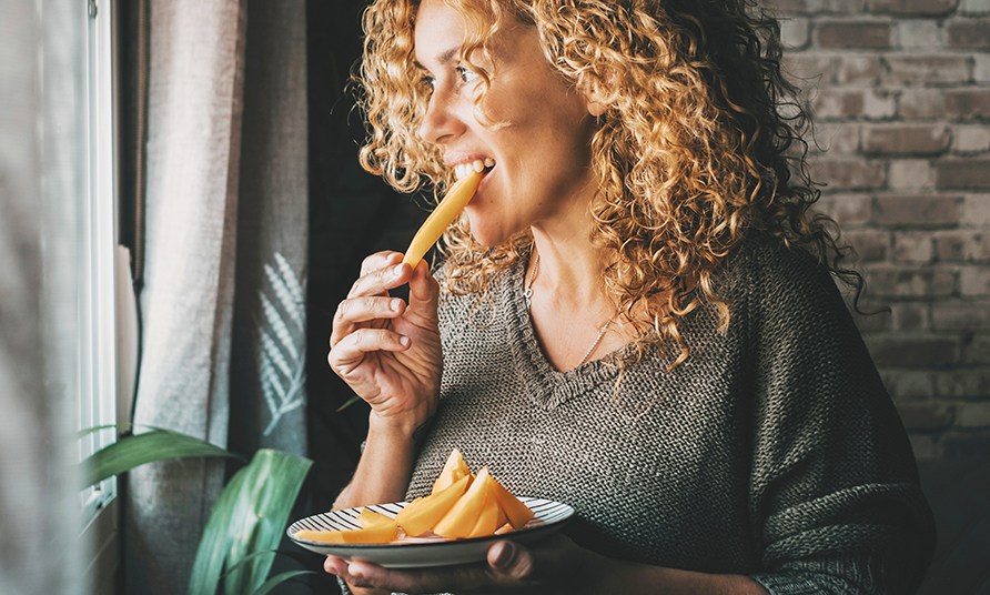 En kvinde st�r ved et vindue og spiser et stykke frugt.