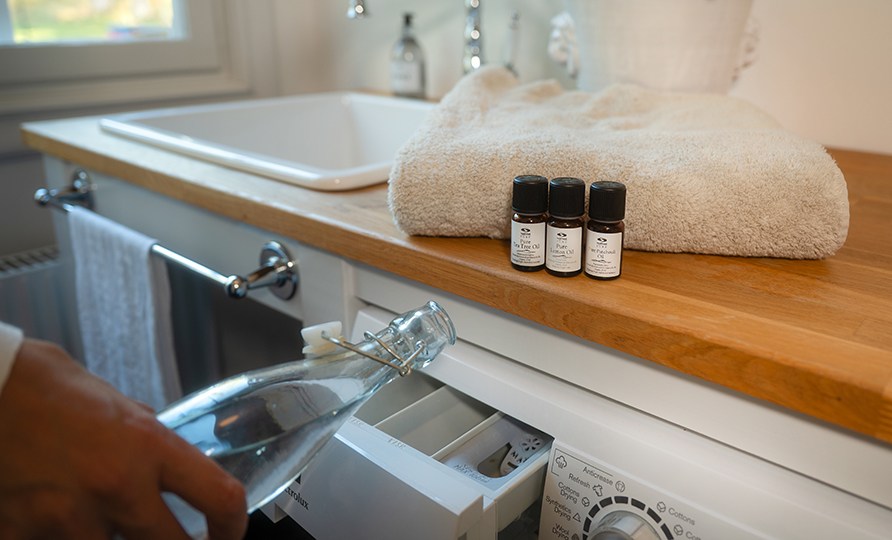 Healthwell PURE æteriske olier står på en vaskemaskine, imens en kvinde hælder hjemmelavet skyllemiddel i vaskemaskinen. Healthwell PURE æteriske olier står på en vaskemaskine, imens en kvinde hælder hjemmelavet skyllemiddel i vaskemaskinen.
